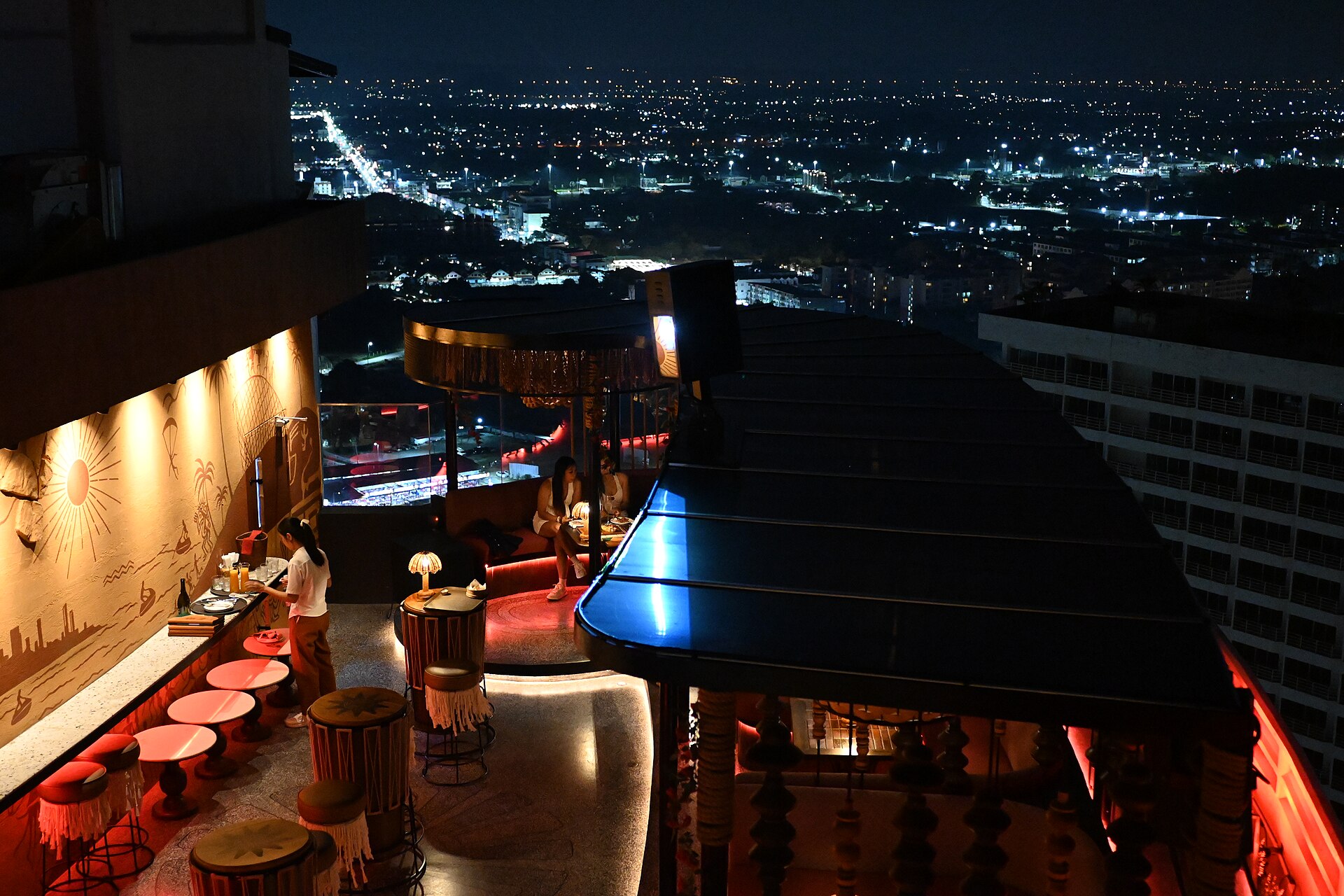 A rooftop bar at night, a staffer tending a counter lined with stools and patrons on a curved banquette, overlooking a wide expanse of city lights below.