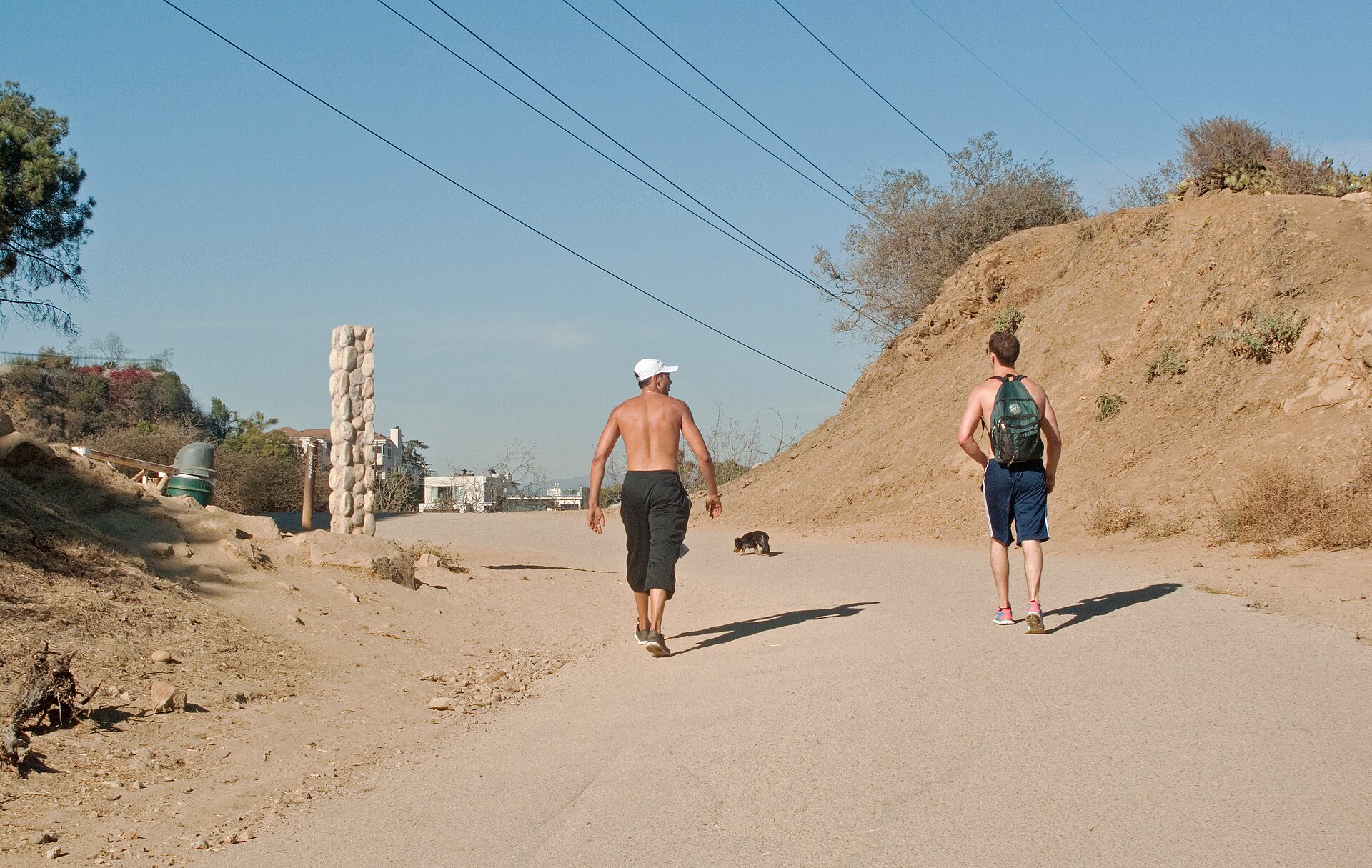 Two shirtless hikers and a small dog climb a wide dirt fire road on a brushy hillside past a stacked-stone pillar under a clear blue sky.