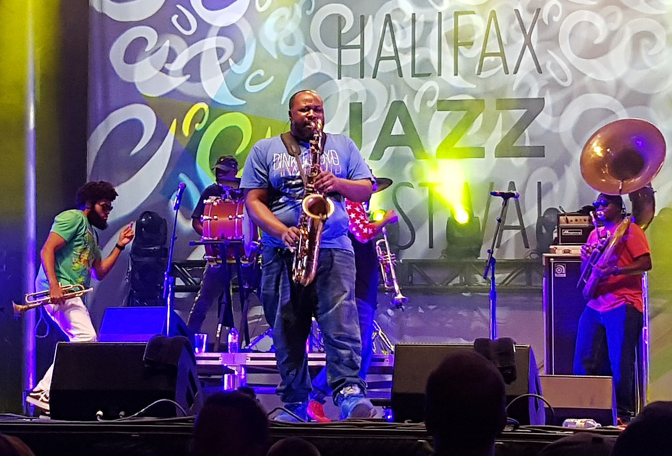 A saxophonist plays center stage under blue light at a jazz festival, backed by a drummer, a trumpeter and a sousaphone player.