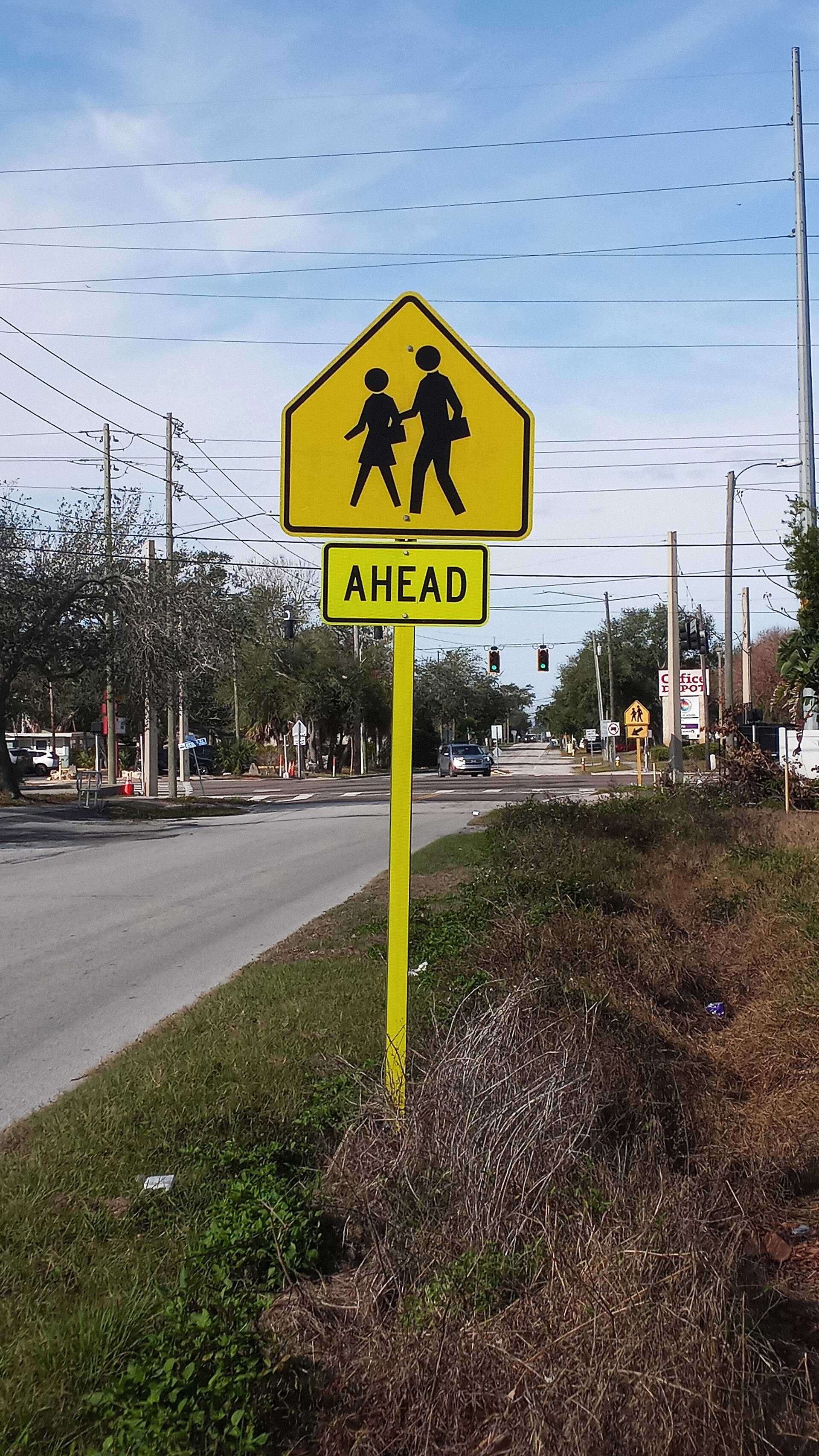 A yellow diamond school-crossing warning sign with an 'AHEAD' plate stands on a yellow post beside a two-lane road, with power lines, distant traffic signals and a strip of dry grass under a blue sky.