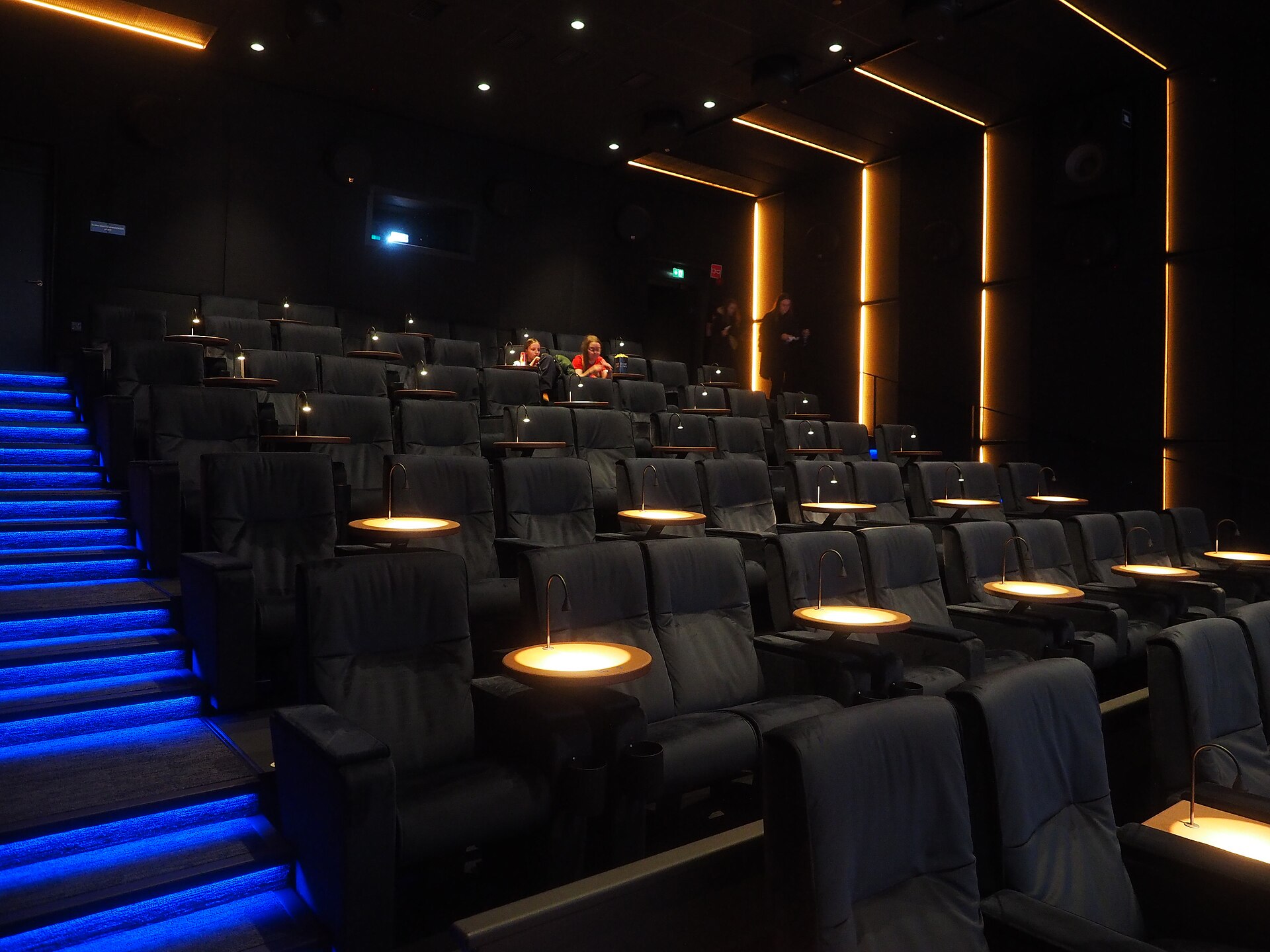 Rows of plush reclining seats, each fitted with a small illuminated lamp, descend toward the front of a darkened modern cinema, blue-lit steps along the left aisle.