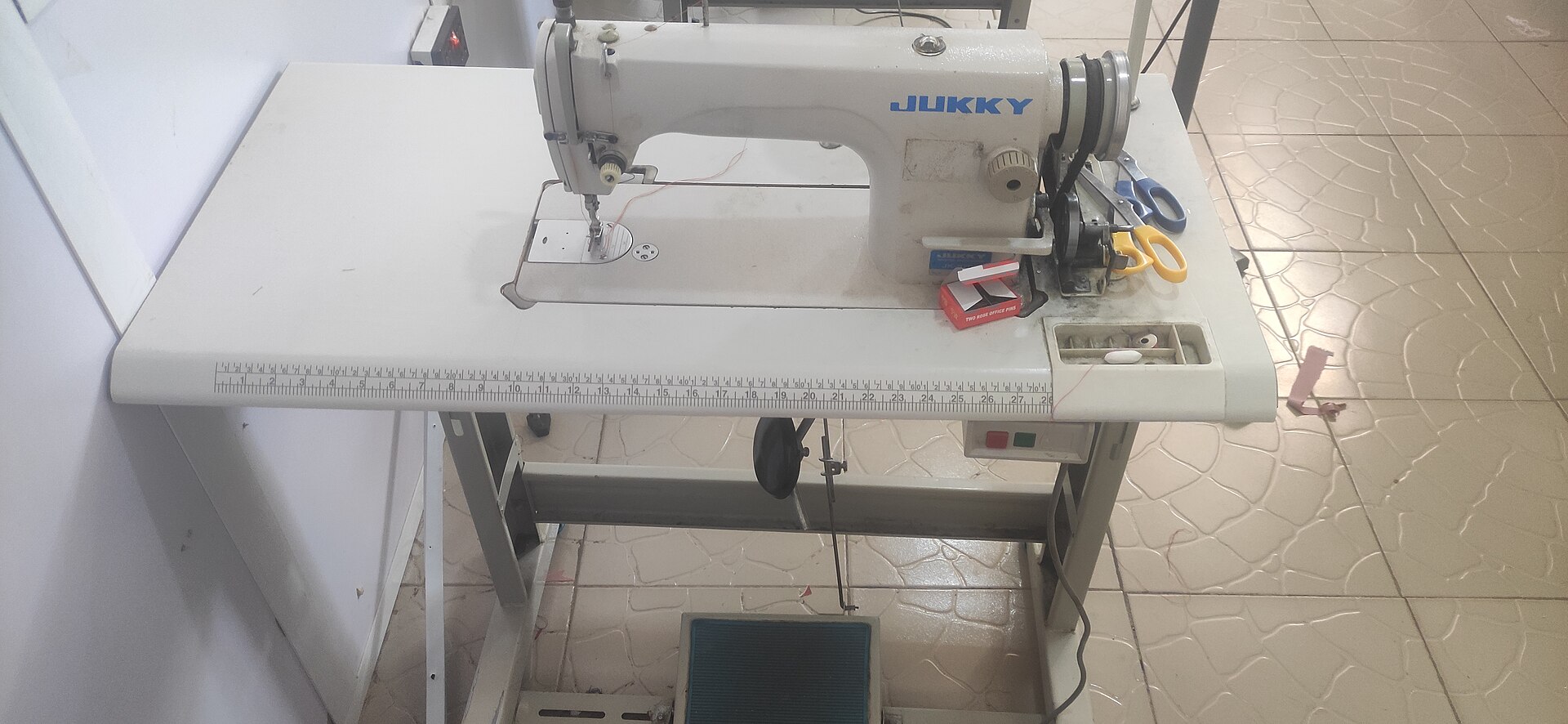 A white industrial sewing machine mounted on a work table with a ruler along its front edge, a pair of scissors and a box of pins beside it.