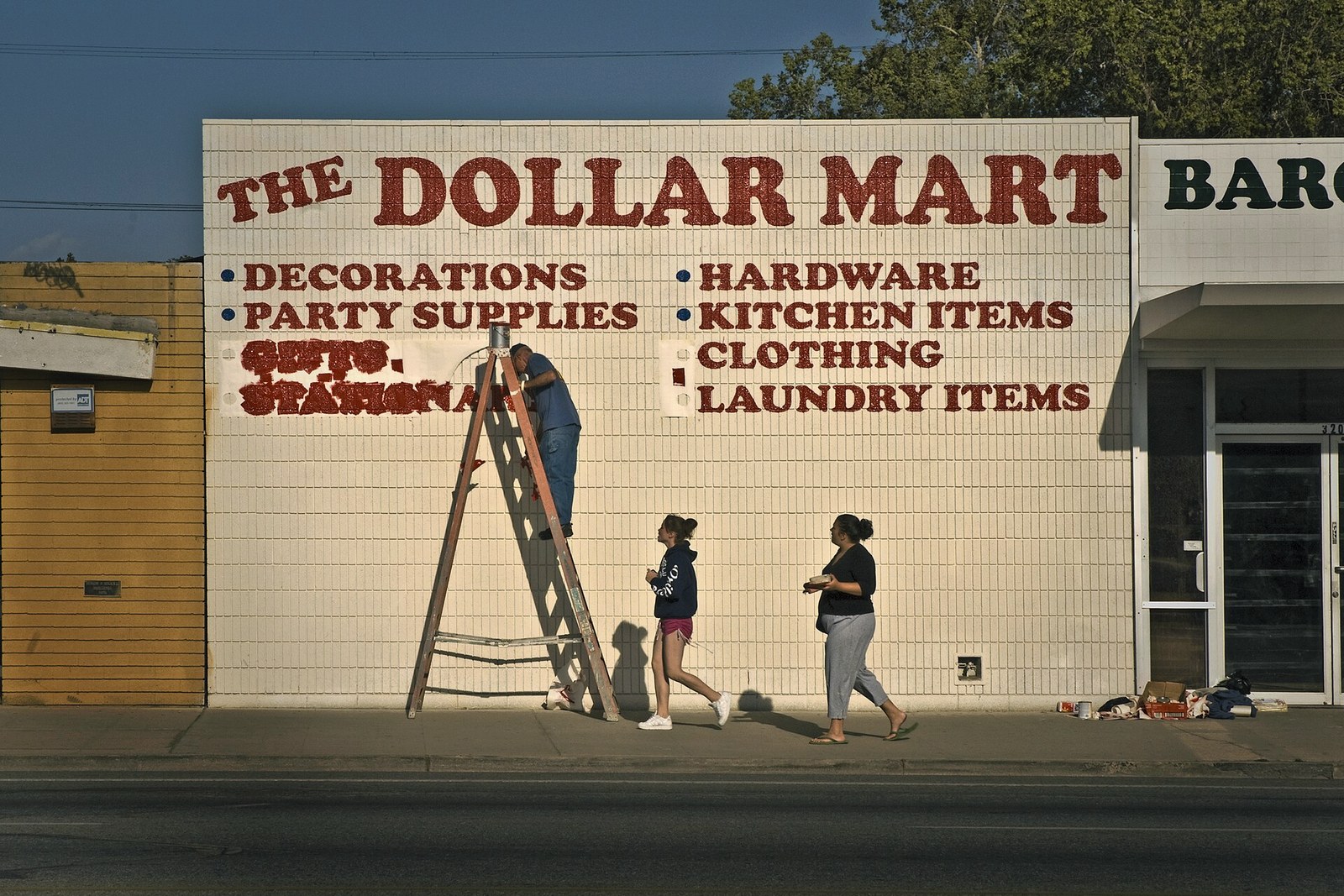 A man in a blue shirt stands on a tall wooden ladder hand-painting red block lettering onto a white tiled storefront wall, while two women walk past on the sidewalk in low evening sun.
