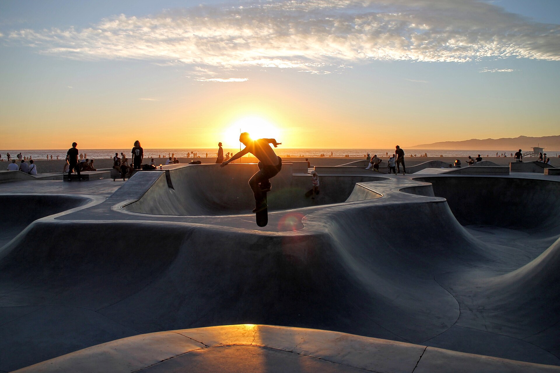 A skateboarder is silhouetted in midair against a low sun above the concrete bowls of a beachside Los Angeles skate park, with onlookers and the ocean behind.