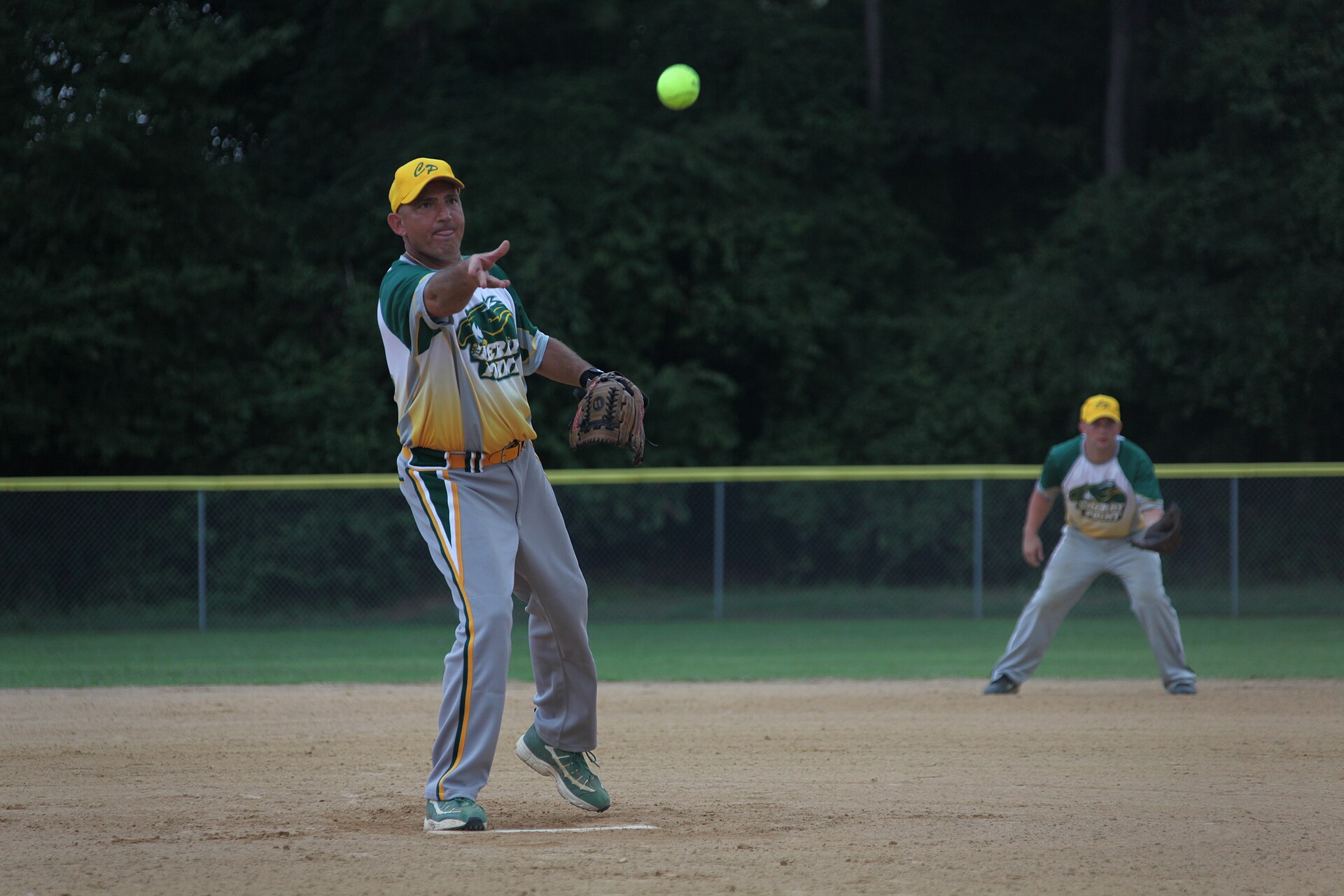An adult player in a green and yellow uniform throws a yellow softball across an infield at dusk as a teammate stands ready behind him.