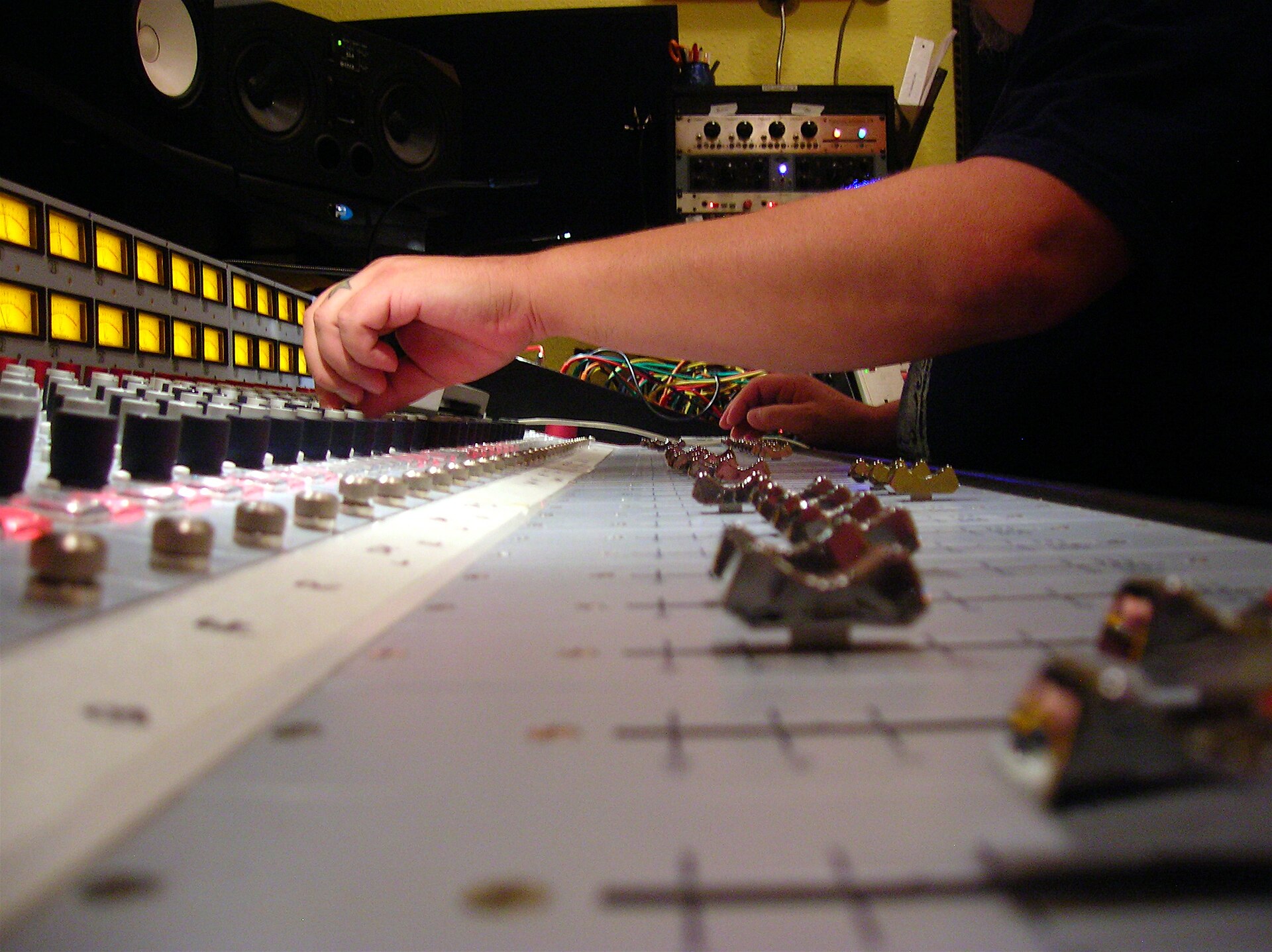 A hand reaches across the faders and glowing meters of a large recording studio mixing console, with studio monitors behind it.