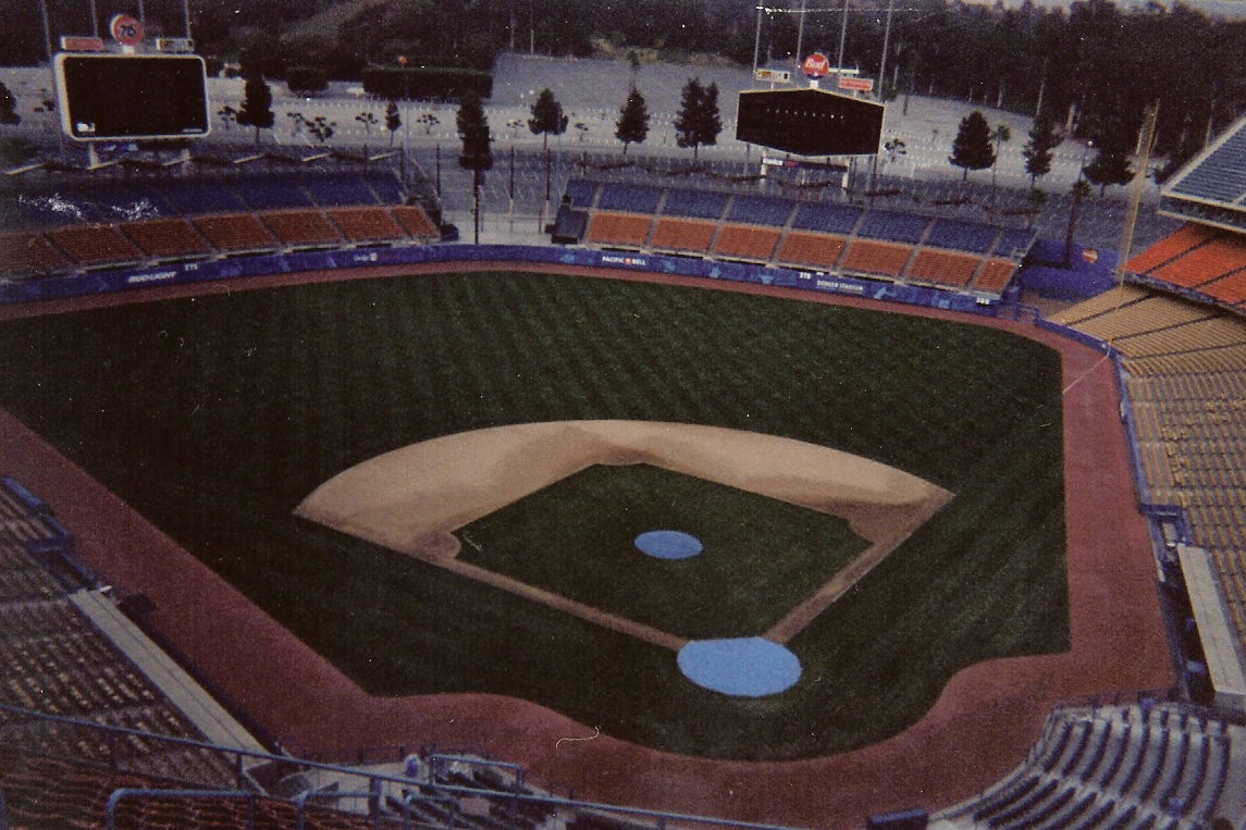 An empty baseball stadium seen from the upper deck at dusk, the field and infield below and tiered seats curving around it, wooded hills behind.