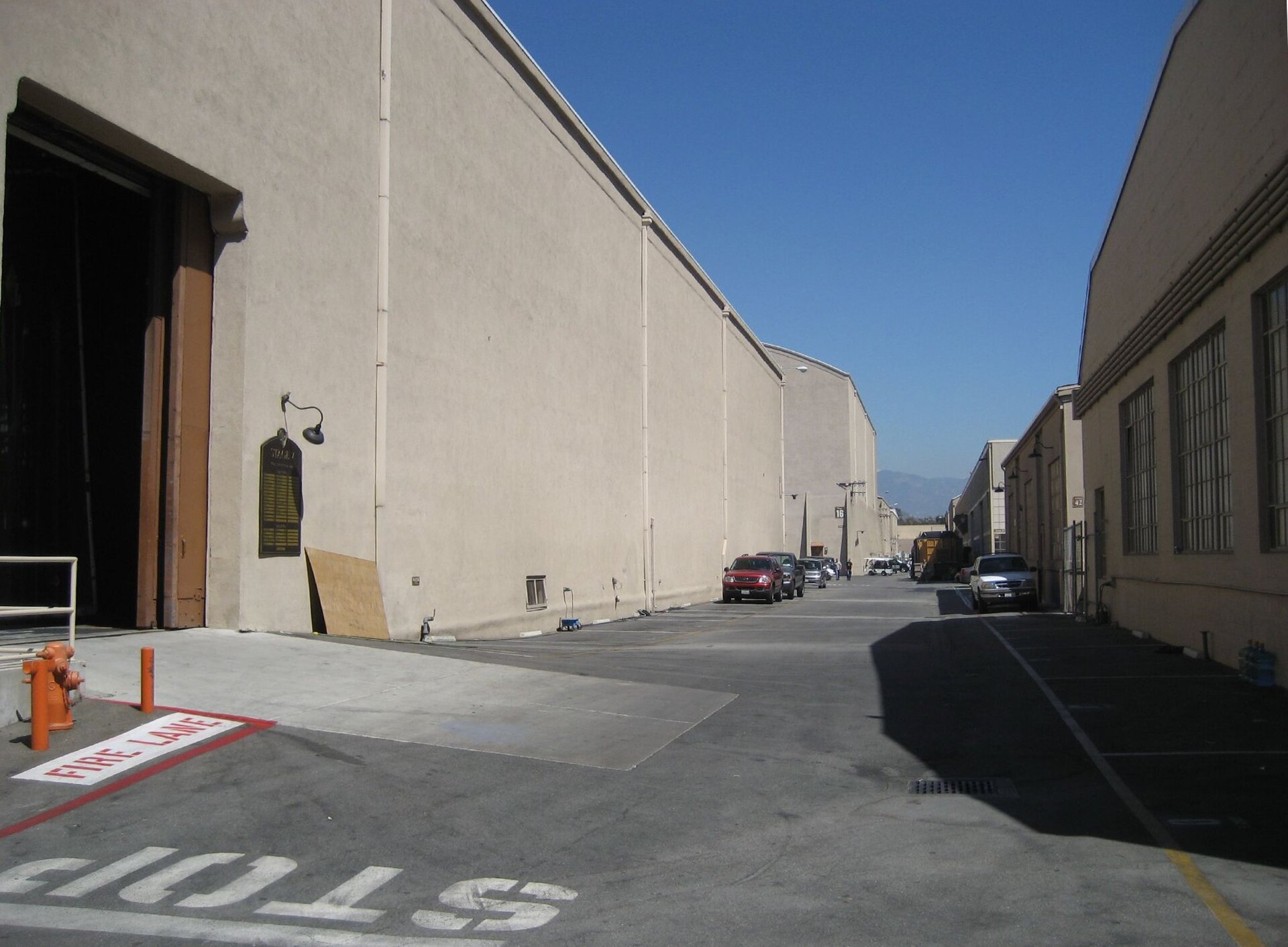 A tall, windowless beige sound stage with its large elephant door standing open at left runs down one side of a studio-lot alley, "FIRE LANE" and "STOP" painted on the pavement, parked cars farther along and mountains in the distance.