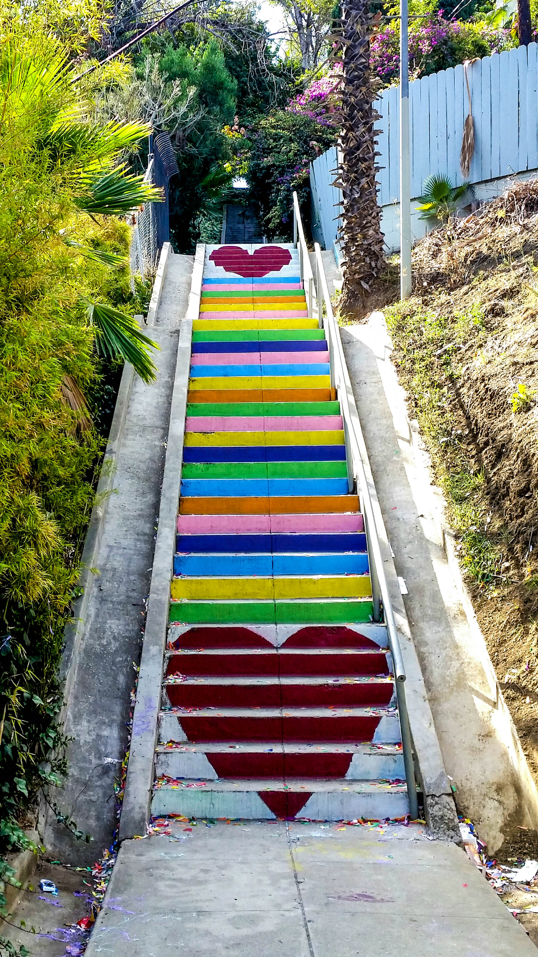 A steep public stairway climbs a hillside, its steps painted in bright rainbow stripes and marked with two large red hearts, flanked by palms, shrubs and a wooden fence.