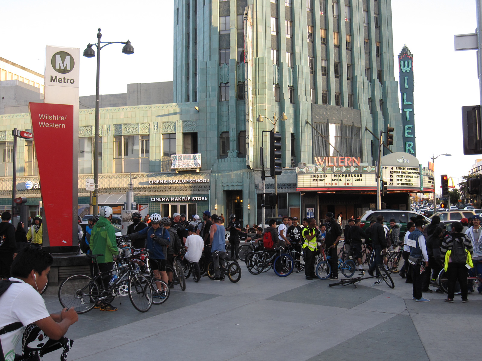 A crowd of cyclists stands with their bicycles on a sidewalk beside a Metro station entrance and an Art Deco theater with a lit vertical marquee, in the fading light of early evening.
