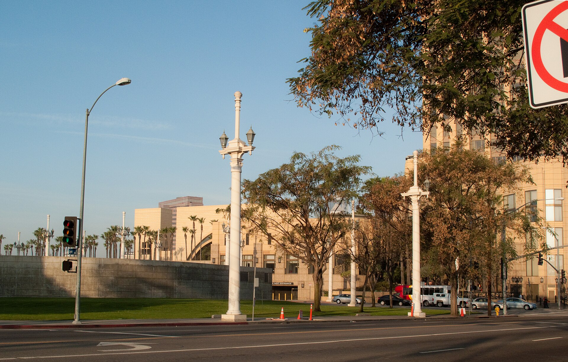 Tall pale ornamental light poles, each topped with a pair of lantern-style lamps, stand along a sunlit street with palm trees and buildings behind.