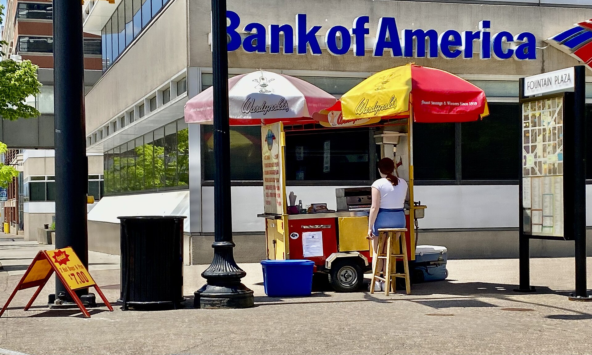 A vendor in a white T-shirt tends a hot-dog cart shaded by red-and-yellow umbrellas on a sunny street corner, a chalkboard price sign and a blue tub on the pavement nearby.