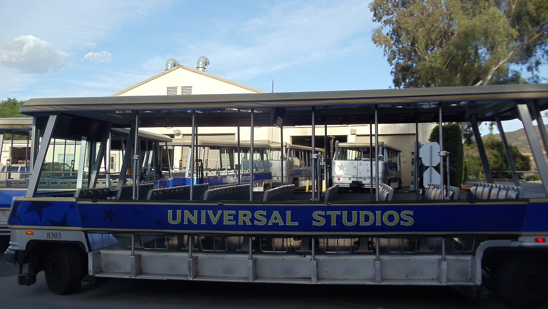 An empty open-sided tour tram lettered "UNIVERSAL STUDIOS" stands parked at a depot, more trams and a building behind under a blue sky.