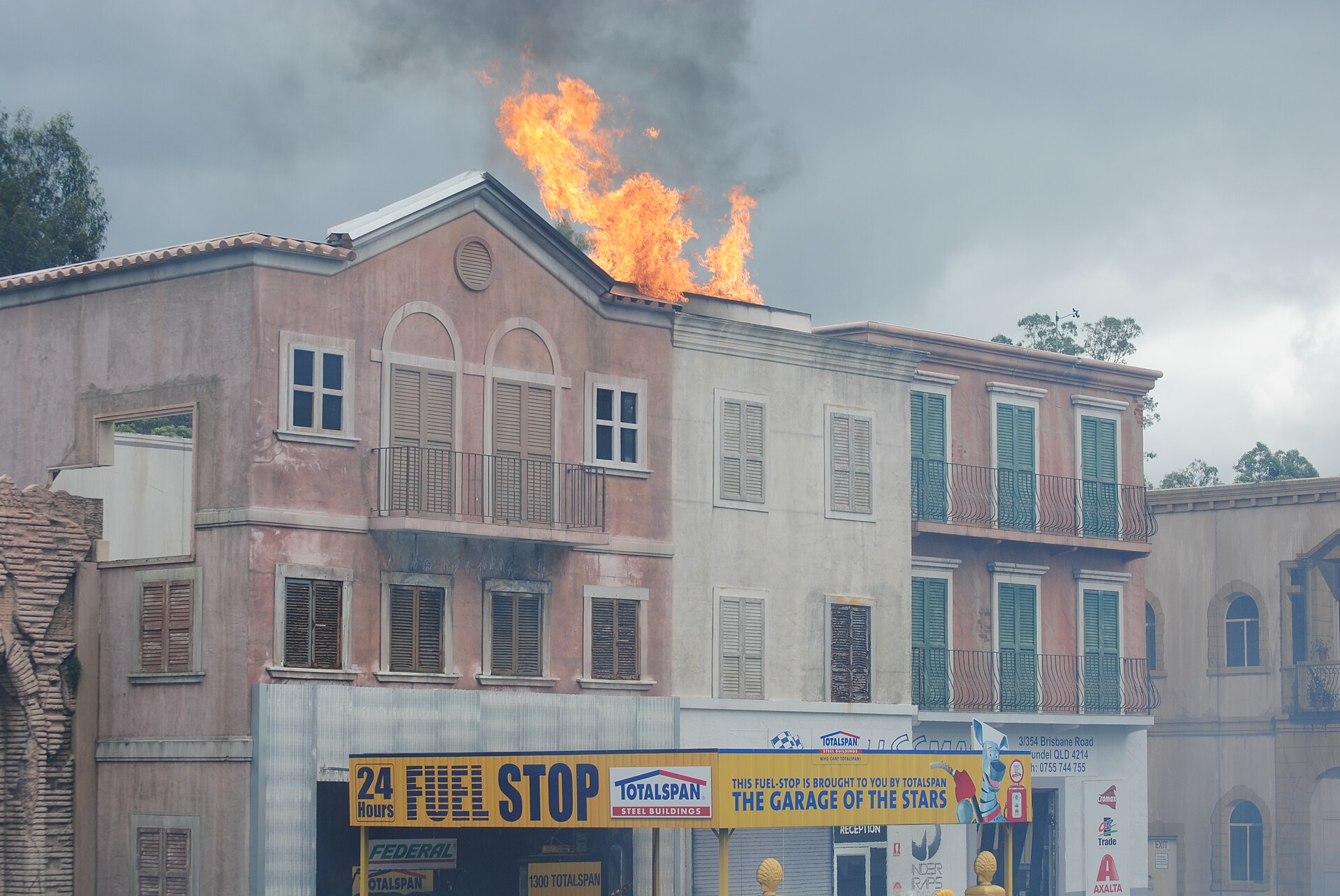 A large fireball and black smoke erupt from the rooftop of a mock building facade during an outdoor stunt show, "FUEL STOP" signage across the storefront below.
