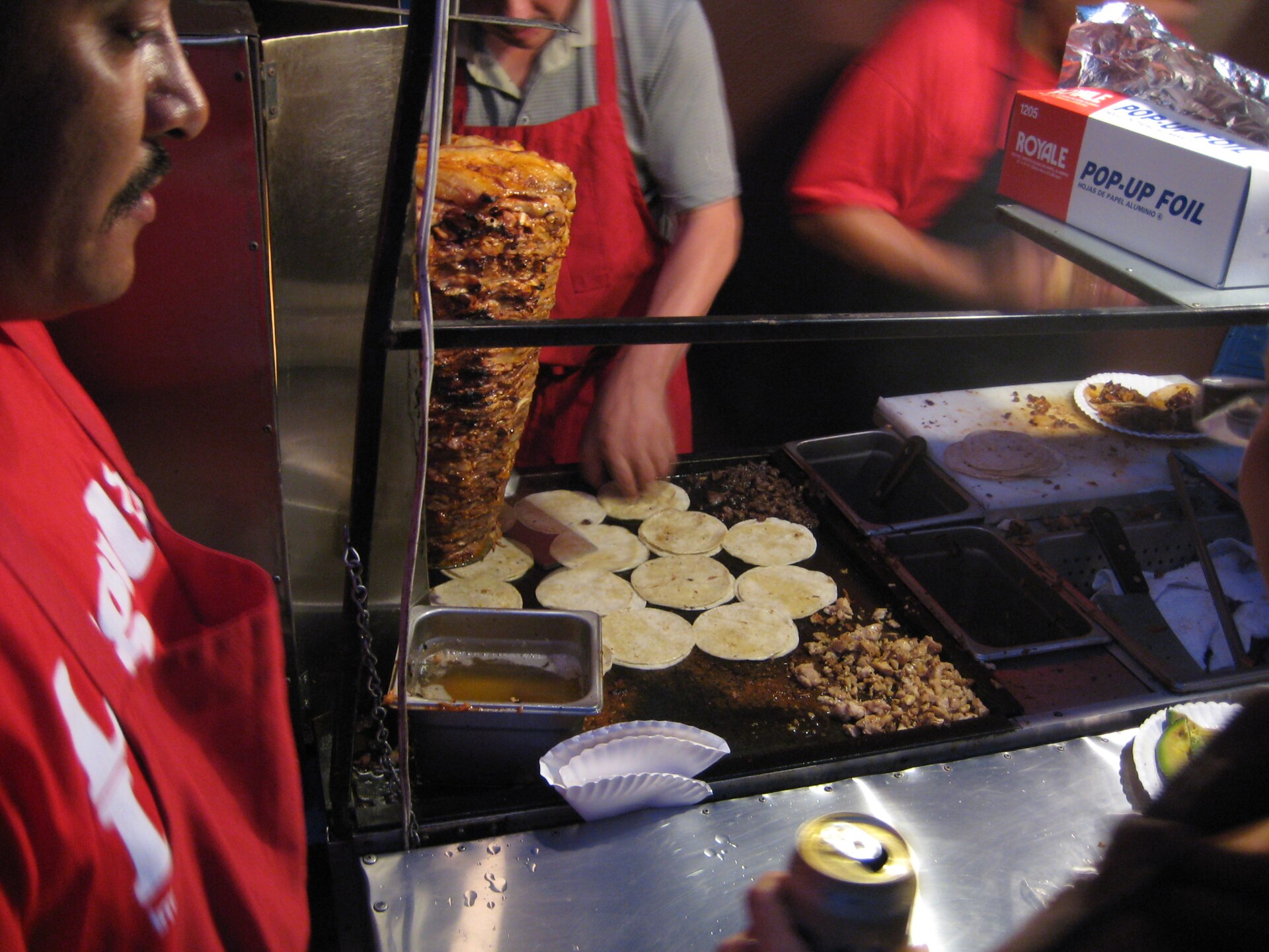 A taco vendor cooks tortillas and chopped meat on a flat griddle beside a vertical spit of al pastor pork at night, workers in red aprons behind the cart.