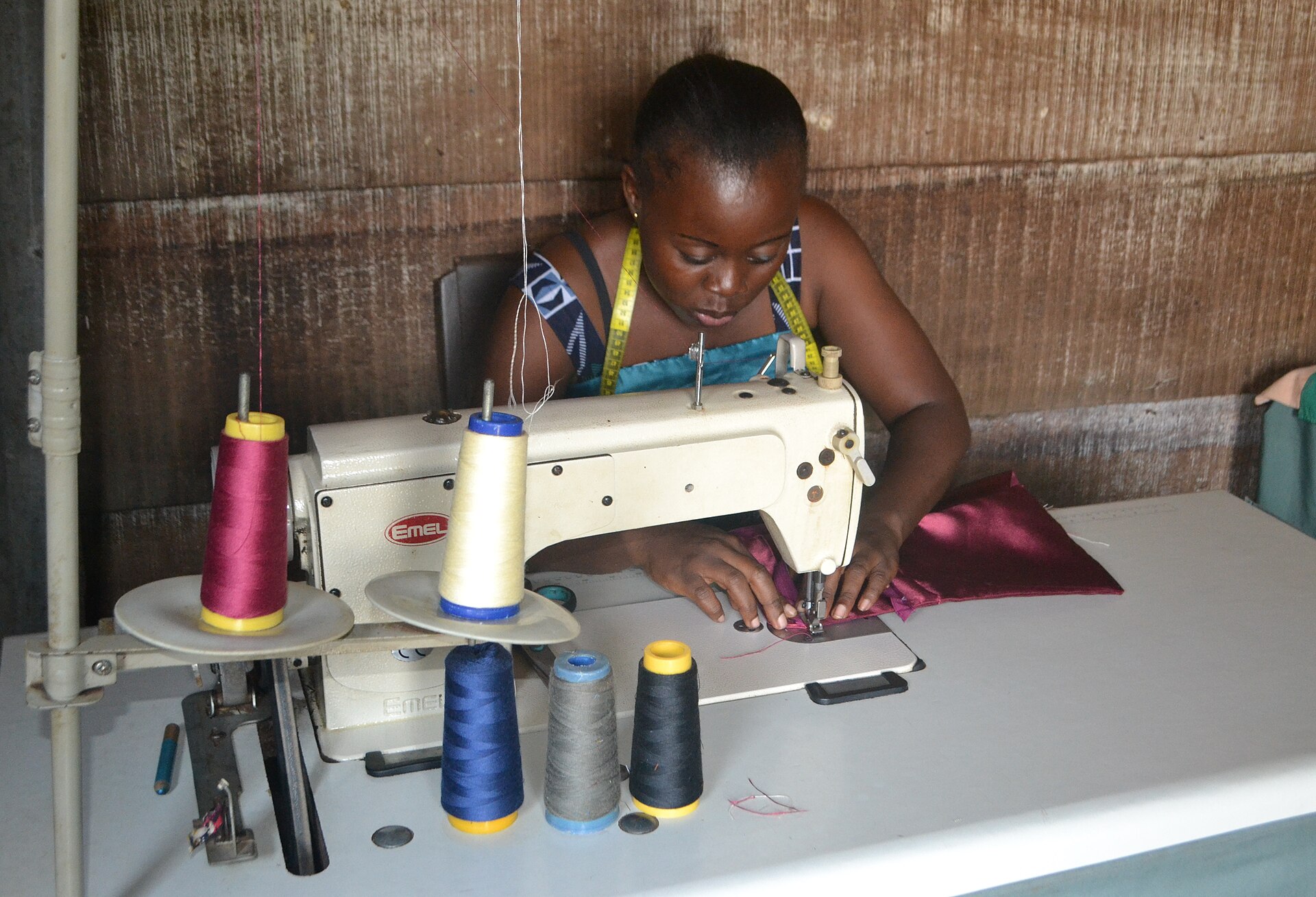 A seamstress with a tape measure around her neck guides bright magenta fabric through an industrial sewing machine, cones of colored thread on the table before her.