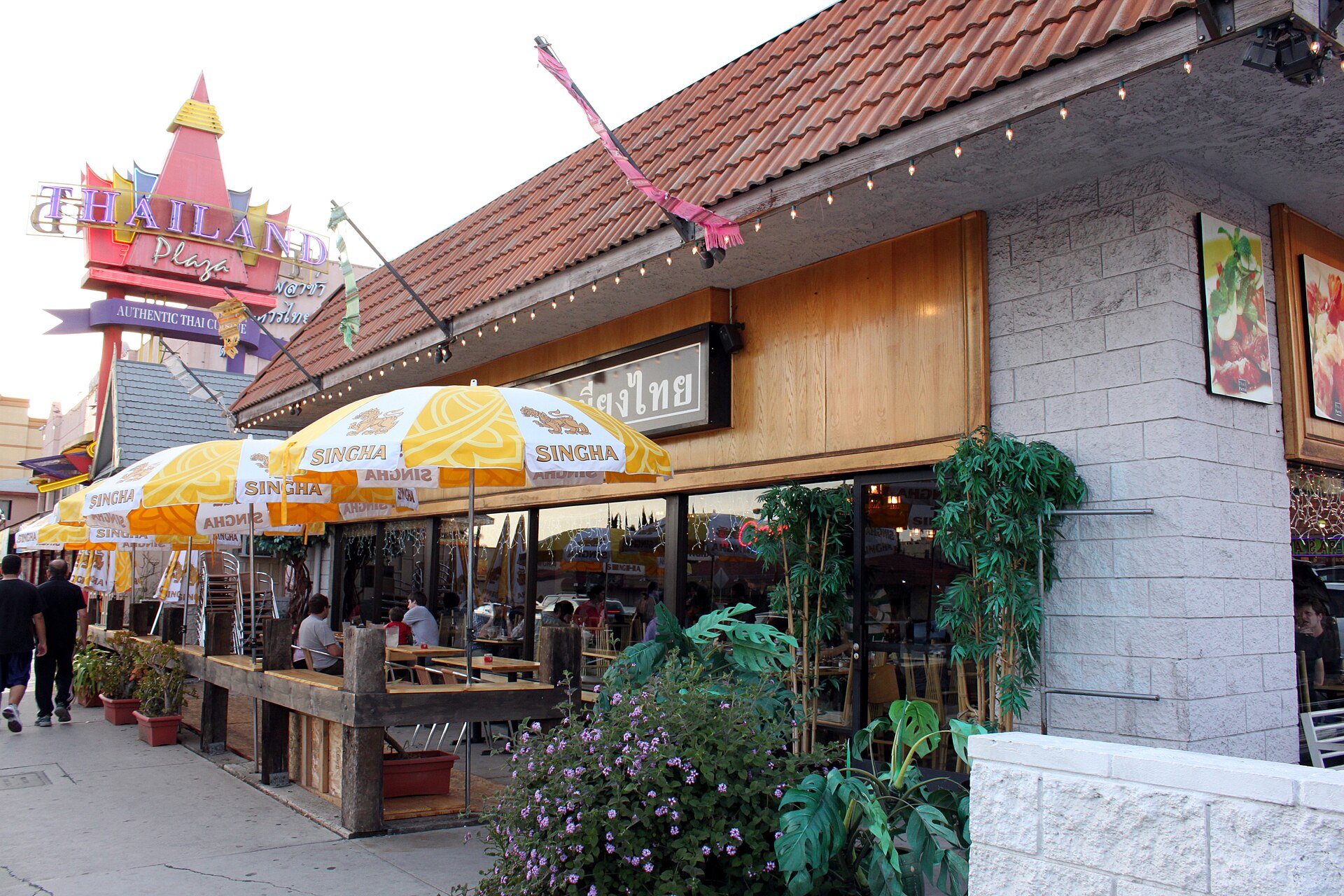 Diners sit at sidewalk tables under yellow Singha-branded umbrellas outside a Thai restaurant with a tiled roof and a tall illuminated sign.