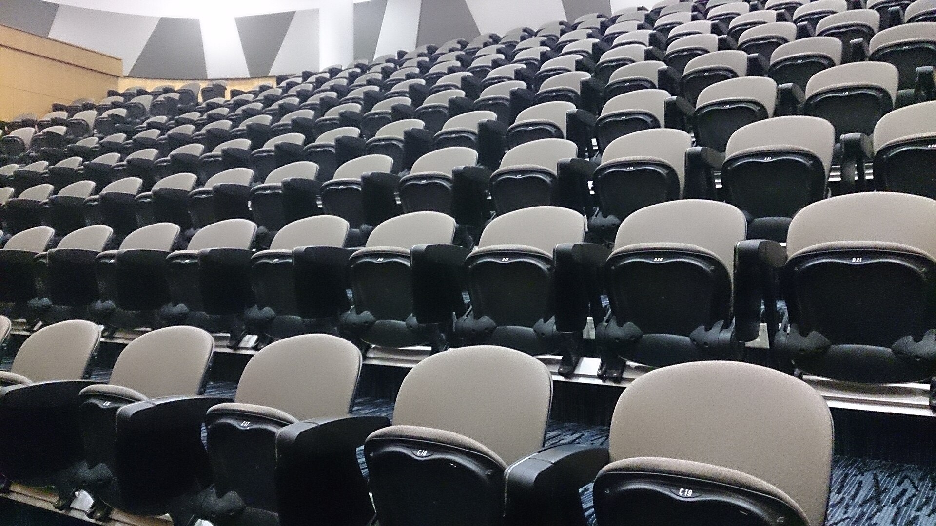Rows of empty upholstered theater seats, alternating gray and black, curve up toward the back of a darkened auditorium, each seat marked with a small row-and-number label.