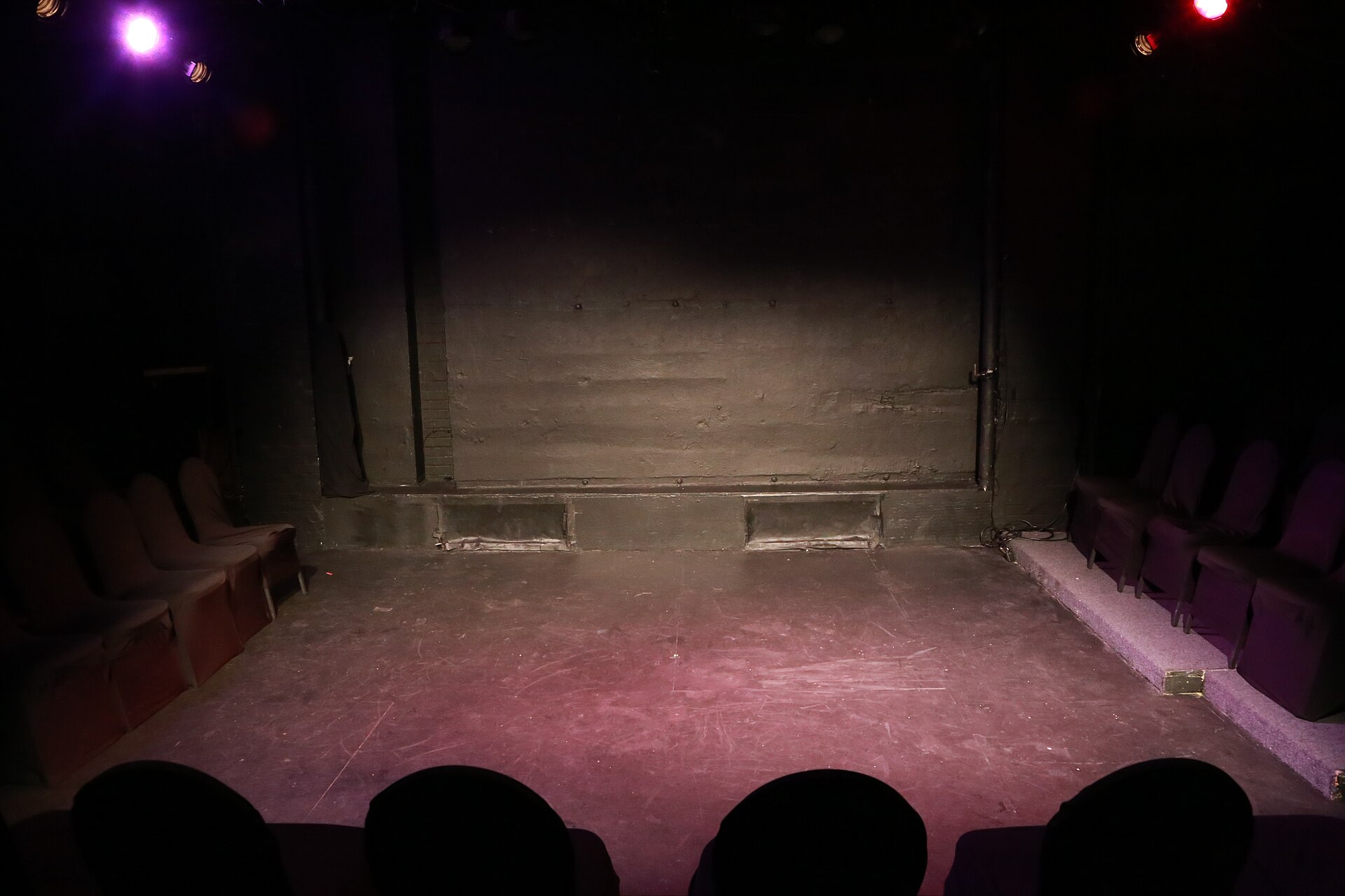 The bare floor of a small black-box theater stage lit in pink and purple, with short rows of empty upholstered seats along either side.