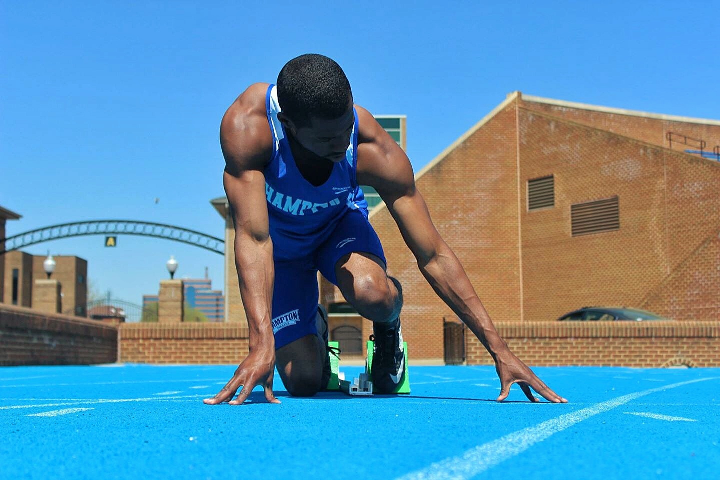 A male sprinter in a blue uniform crouches in the set position in starting blocks on a blue track, a brick building behind under a clear sky.