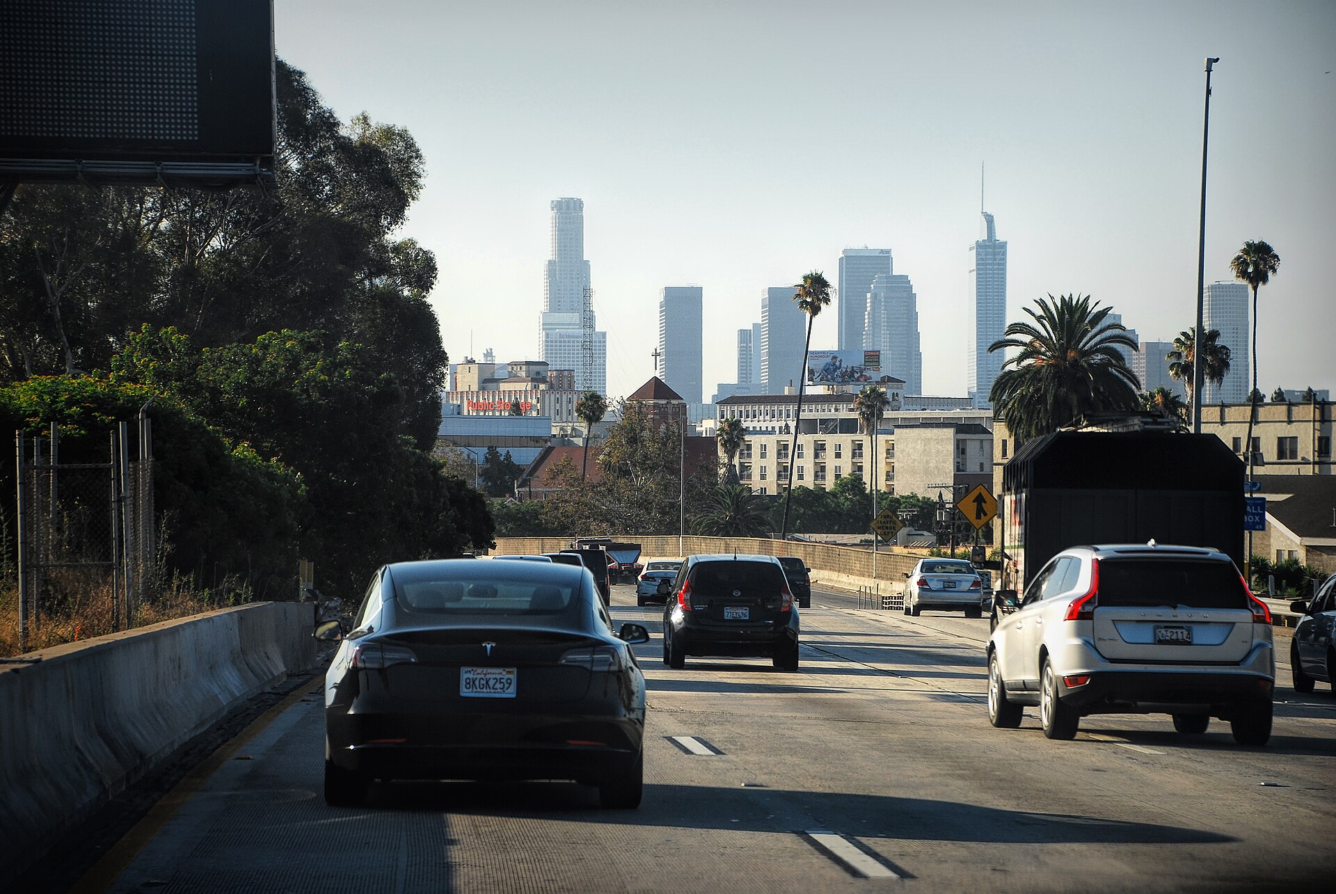 Cars fill the lanes of a Los Angeles freeway in afternoon light, the downtown skyline hazy in the distance beyond palm trees.