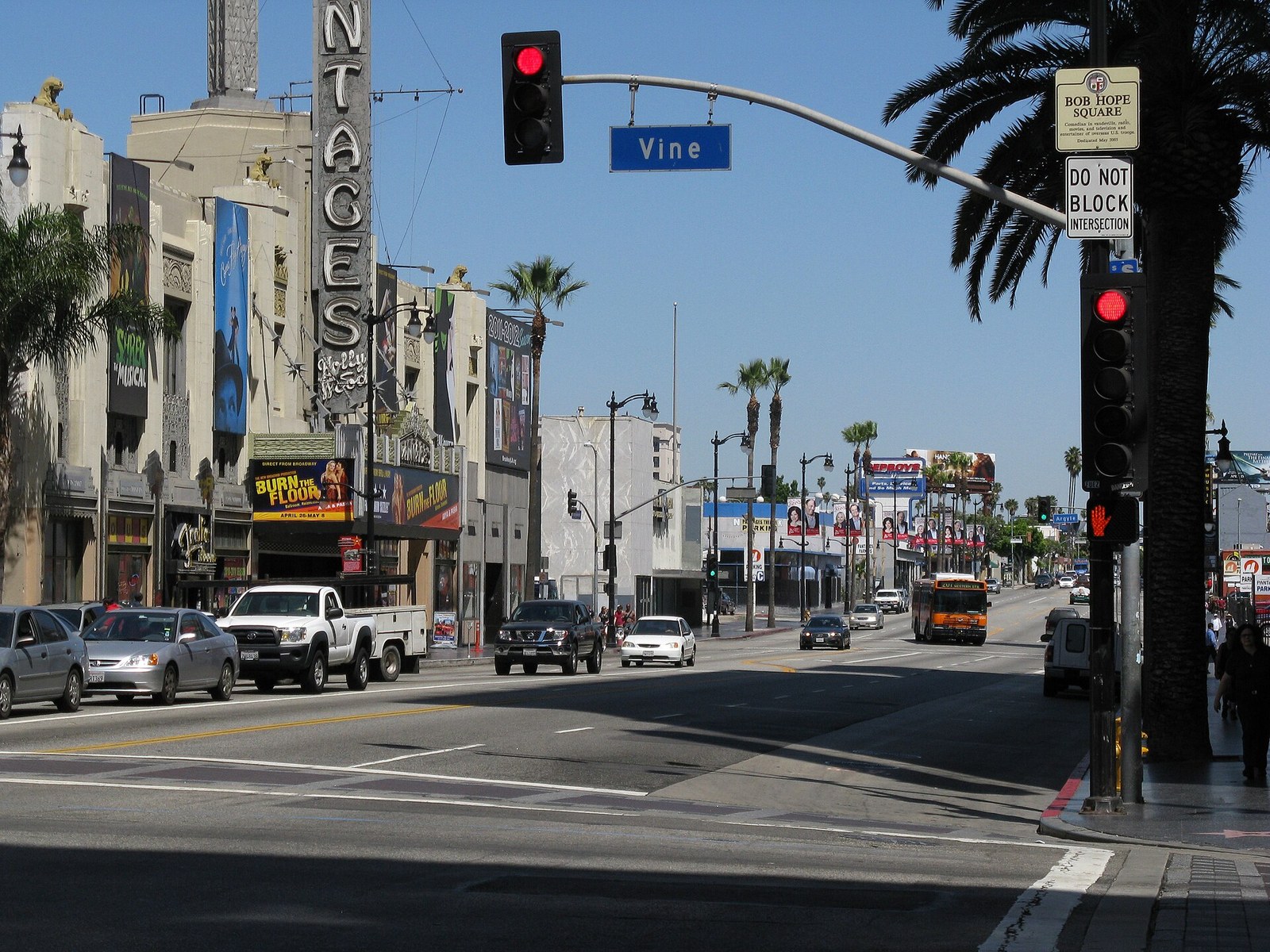 Looking west along Hollywood Boulevard at Vine Street: a blue "Vine" street sign hangs from a traffic-signal mast beside a "Bob Hope Square" plaque, with a theater marquee, palm trees, cars and a bus along the street.