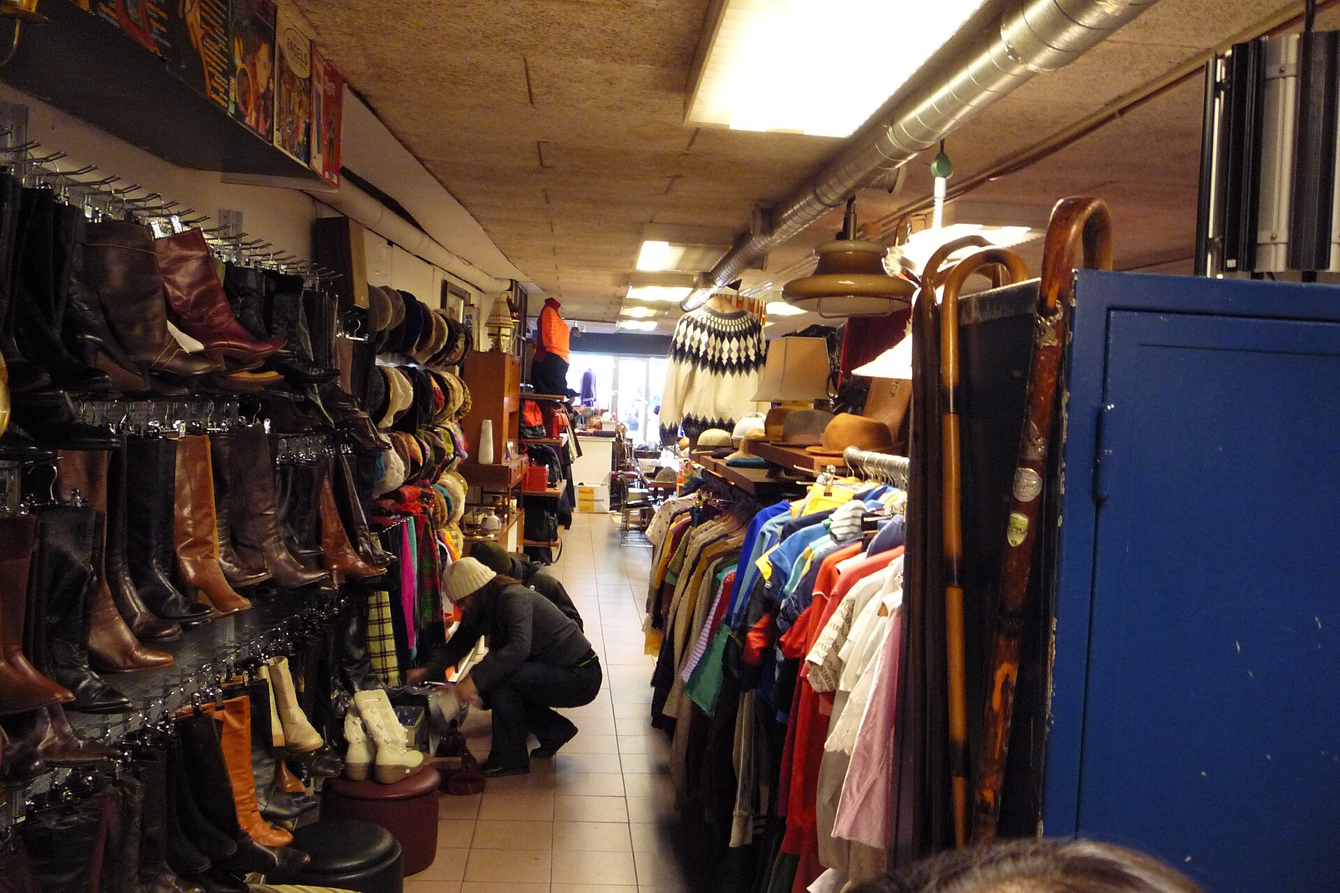 A crowded secondhand shop with a wall of leather boots on the left, racks of assorted clothing on the right, hats and lamps on shelves, a customer crouching to look through items.