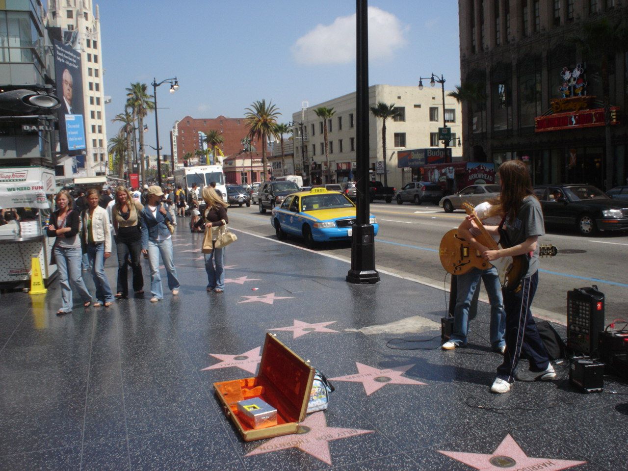 A guitarist plays on the terrazzo stars of the Hollywood Walk of Fame, an open guitar case and small amplifiers at his feet, as pedestrians pass and a taxi drives by in bright daylight.